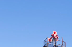 Worker in red safety jacket on a high metal scaffold against a clear blue sky with a hard hat on top platform edgey.