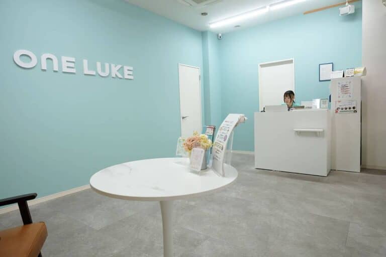 Reception area with teal walls and a white front desk; 'ONE LUKE' sign on the wall, and a round table with flowers and brochures in the foreground.